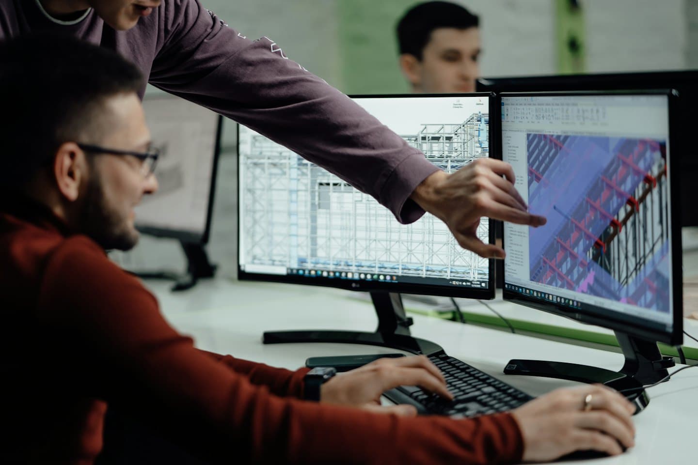 Un uomo indica un progetto 3D su doppio monitor mentre un collega digita.